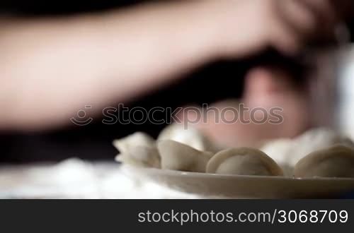 Focus on the plate with raw pelmeni, then on hands making them, pinching the dough closed and forming it