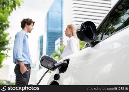 Focus parking-electric car connected to public charging station with blur progressive businesspeople holding coffee, residential building apartment and condo background for eco-friendly concept.. Progressive businesspeople with coffee and EV car at public charging station.