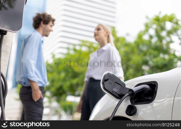Focus parking-electric car connected to public charging station with blur progressive businesspeople holding coffee, residential building apartment and condo background for eco-friendly concept.. Progressive businesspeople with coffee and EV car at public charging station.
