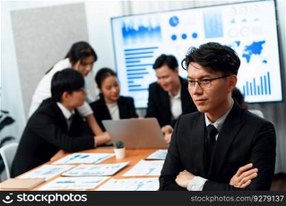 Focus portrait of successful confident male manager or executive in business wear with blurred background of businesspeople, colleagues working with financial report papers in office of harmony.. Focus portrait male manager in harmony office with businesspeople in background.