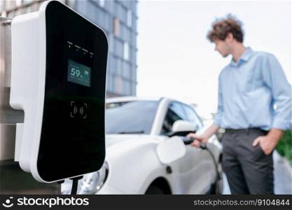 Focus public EV charging station in city residential area with blur background of businessman charging electric vehicle. Progressive lifestyle of ecological concern of city-dwelling individual.. Progressive concept of focus EV car at charging station with blur man background