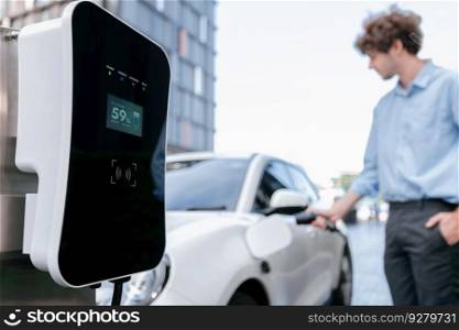 Focus public EV charging station in city residential area with blur background of businessman charging electric vehicle. Progressive lifestyle of ecological concern of city-dwelling individual.. Progressive concept of focus EV car at charging station with blur man background