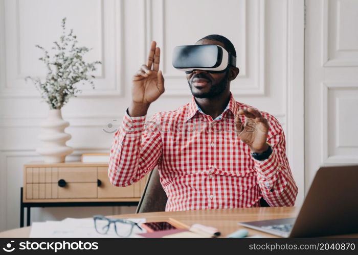 Focused Aframerican man in VR glasses enjoying augmented reality while sitting at workplace at home office, pointing with fingers up in air interacting with virtual 3D objects, using electronic device. Focused Aframerican man in VR glasses enjoying augmented reality while sitting at workplace