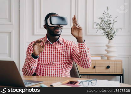 Focused Aframerican man in VR glasses enjoying augmented reality while sitting at workplace at home office, pointing with fingers up in air interacting with virtual 3D objects, using electronic device. Focused Aframerican man in VR glasses enjoying augmented reality while sitting at workplace