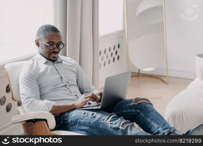 Focused african american man freelancer working online on project, sitting in armchair with laptop. Modern black businessman wearing glasses typing message, answering email. Remote job concept.. African american man freelancer works online on project sitting in armchair with laptop. Remote job