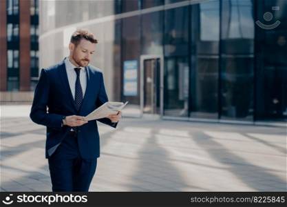 Focused bearded young businessman in formal outfit walking alone with newspaper in hands, reading latest news on his way to workplace with office buildings in background. Focused formally dressed business person reading newspaper while walking near office building