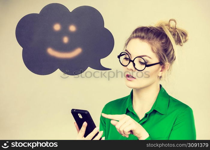 Focused business woman wearing green shirt and red eyeglasses looking at phone with black thinking or speech bubble next to her.. Focused business woman looking at phone, thinking bubble