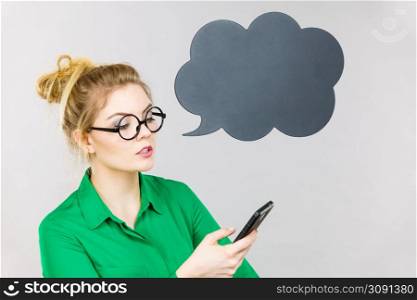 Focused business woman wearing green shirt and red eyeglasses looking at phone with black thinking or speech bubble next to her.. Focused business woman looking at phone, thinking bubble