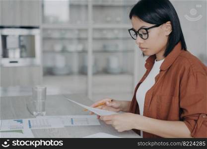 Focused businesswoman analyzing documents with statistics, working on financial project on workspace. Female employee wearing glasses prepares a report. Strategic planning, data analysis.. Focused businesswoman analyzing documents with statistics, working on financial project on workspace