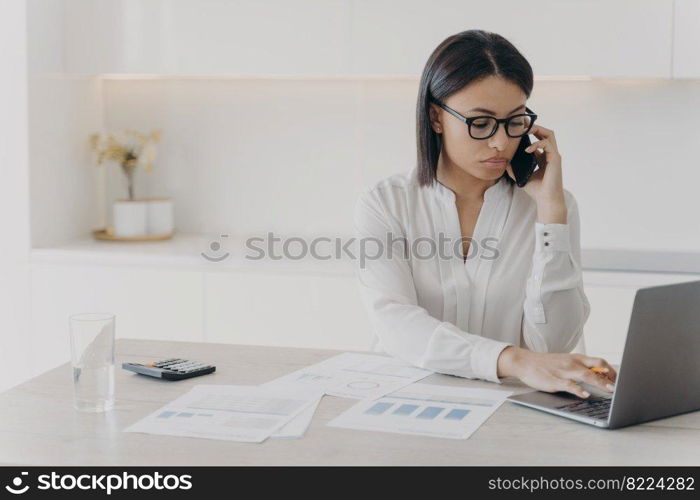 Focused businesswoman answers mobile call, consulting client, working on laptop at office desk. Serious female manager wearing glasses communicates with coworker, typing on computer.. Focused businesswoman answers mobile call, consulting client, working on laptop at office desk