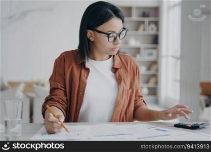 Focused businesswoman in glasses calculates expenses, plans company budget with a calculator. Dedicated female accountant freelancer works on financial reports. Effective financial management.