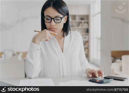 Focused businesswoman in glasses counts profit on calculator, analyzes company budget. Female manager working with documents, calculates expenses, sitting at office desk. Financial management.. Businesswoman in glasses counts profit on calculator, analyzes company budget. Financial management