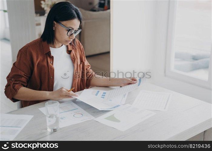 Focused businesswoman in glasses works on project, prepares report in charts analyzing documents at home office. Serious woman manager analyzes graphs, looking through papers on workplace.. Focused businesswoman works on project, prepares report in charts analyzing documents at home office
