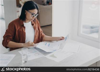 Focused businesswoman in glasses works on project, prepares report in charts analyzing documents at home office. Serious woman manager analyzes graphs, looking through papers on workplace.. Focused businesswoman works on project, prepares report in charts analyzing documents at home office