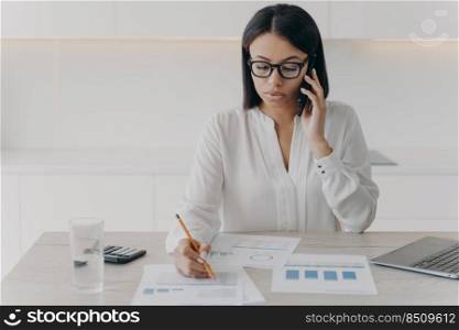 Focused businesswoman talking on phone, discussing project, financial analytics. Serious female businessperson having call, communicates with client writing note, sitting at office desk.. Focused businesswoman talk on phone, discuss project, financial analytics, write note at office desk