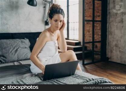 Focused businesswoman wrapped in towel after bathing working at laptop sitting on bed in the morning. Female freelancer checking work email on computer at home. Remote job concept.. Focused businesswoman works at laptop sitting on bed after shower in the morning at home. Remote job
