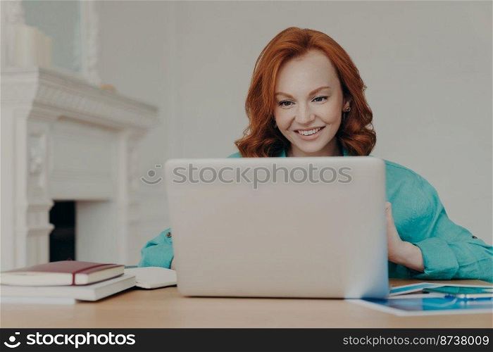 Focused cheerful redhead successful businesswoman does research, makes business project, sits in front of laptop computer at home office desk, uses software application, has happy smile on face