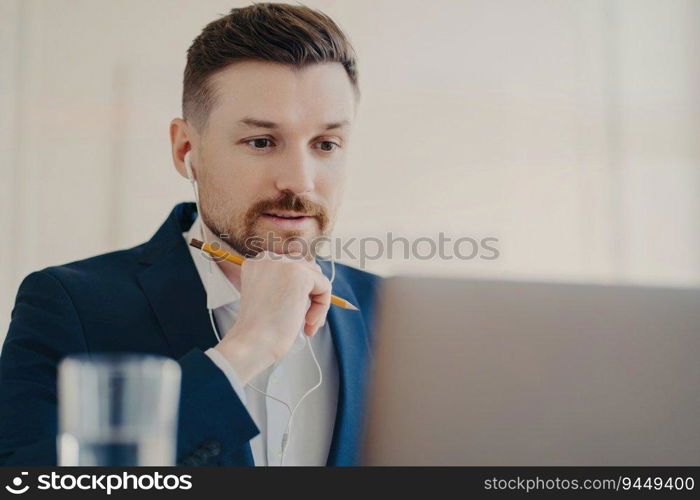 Focused entrepreneur in suit, earphones on, multitasking online with laptop, pencil, and webinar.