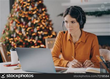 Focused European lady support consultant in headset with mic working at christmas time from home, pensive female freelancer using laptop while sitting in room with sparkling Xmas tree on background. Focused European lady support consultant in headset with mic working at christmas time from home