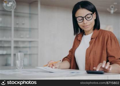 Focused female employee wearing glasses counts expenses on calculator, planning financial project budget. Pleased businesswoman calculates profit, analyzing documents, sitting at office desk.. Female employee in glasses counts expenses on calculator, planning financial project budget at desk