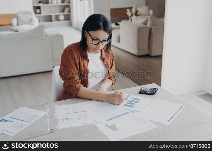 Focused female freelancer analyzing financial statistics, calculates expenses, works on project at home. Businesswoman managing company budget, working, sitting at desk full of documents.. Female freelancer analyzing financial documents, calculates expenses, works on project at home