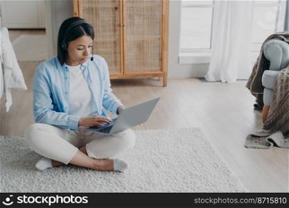 Focused female freelancer wearing headset works on laptop, sitting on the floor at home. Serious woman in wireless headphones watching educational webinar, studying online. E-learning, remote job.. Female wearing headset works on laptop typing, sitting on the floor at home. E-learning, remote job