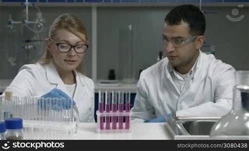 Focused female supervisor in protecive gloves and her mixed race male assistant scientist pipetting solution into test tubes in biochemistry laboratory. Concetrated scientific researcher analyzing samples during chemical experiment in lab.