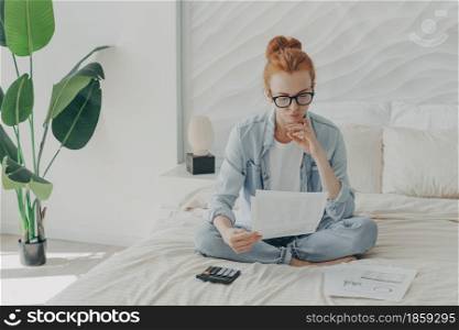 Focused ginger european woman sitting on bed and calculating domestic bills at home, holding papers, reading bank notification while accounting monthly budget and planning family expenses. Focused ginger european woman sitting on bed and calculating domestic bills at home, holding papers