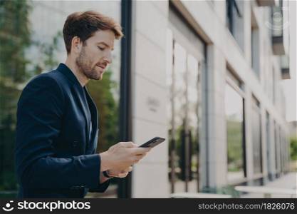 Focused handsome man with beard wearing dark suit chatting on smartphone, browsing web on phone while standing in front of building with lots of windows, selective focus on businessman typing message. Handsome young man chatting and browsing web on his phone while standing outdoors