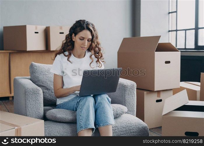 Focused hispanic girl works at laptop searching ideas for renovation, choosing furniture, shopping interior decor for new house online, sitting on armchair among card boxes. Moving, relocation concept. Girl shopping online for new house at laptop, sitting among card boxes. Relocation, ecommerce