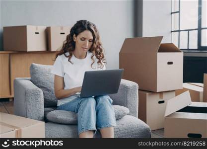 Focused hispanic girl works at laptop searching ideas for renovation, choosing furniture, shopping interior decor for new house online, sitting on armchair among card boxes. Moving, relocation concept. Girl shopping online for new house at laptop, sitting among card boxes. Relocation, ecommerce