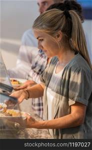 Focused looking attractive woman serves herself food with stainless steel tongs at a cafeteria. Eating concept.. Focused looking woman serving herself food at a cafeteria