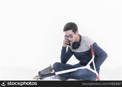 Focused looking teenage boy with headphones hanging on his neck and a scooter resting on his lap sits cross-legged while talking on his phone on a light background. Leisure concept.. Teenage boy talks on his phone while resting
