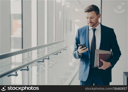 Focused male business consultant dressed formally holding mobile phone, reading news online, standing in office corridor during coffee break or after meeting with collegues. People and technology. Focused business consultant dressed formally holding mobile phone, reading news online in office