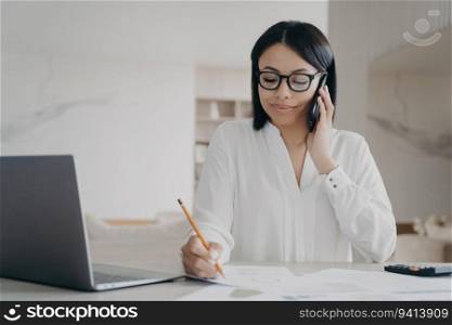 Focused manager answers call, consults client, makes notes at laptop. Businesswoman chats on cellphone at office desk.