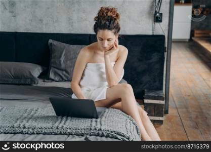 Focused spanish woman wrapped in towel after shower working on laptop sitting on bed. Pensive female remote worker freelancer doing job, checking email in her bedroom in the morning. Distant work.. Focused woman wrapped in towel after shower works on laptop sitting on bed in morning. Distant work