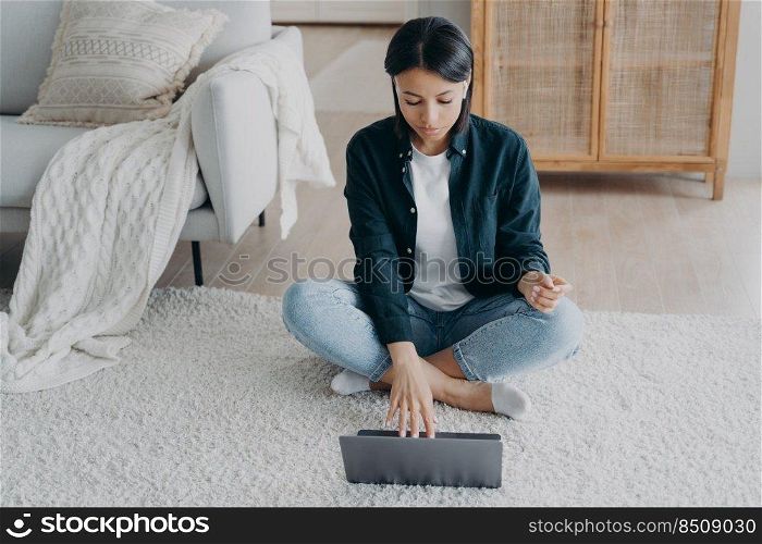 Focused woman working on laptop or learning online sitting on floor in living room at home. Concentrated young female freelance student looking at computer screen, typing. E-learning, remote work.. Focused woman working on laptop or learning online sitting on floor at home. E-learning, remote work