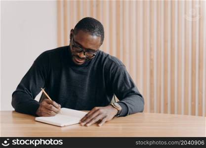 Focused young african race man in glasses taking notes in notebook. Concentrated African ethnicity student learning hard to get highest score and pass exams successfully. Freelance and education concept. Focused young african man in glasses holding pen, taking notes in agenda while sitting at desk