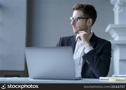 Focused young German male office worker sits at desk at home workplace distracted from laptop screen, pensive businessman looking aside immersed in thoughts of upcoming online meeting. Focused young German male office worker sits at desk at home workplace distracted from laptop screen