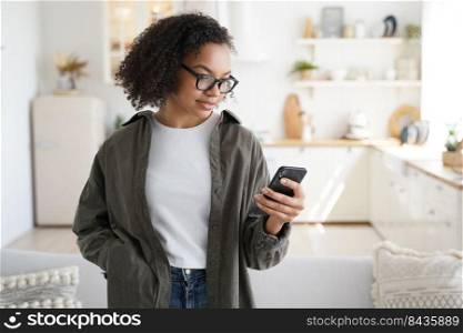 Focused young girl using smartphone at home, shopping groceries for kitchen in online store with mobile apps. Modern female in glasses chatting in social network, browsing content.. Focused young girl using smartphone at home, shopping groceries in online store with mobile apps