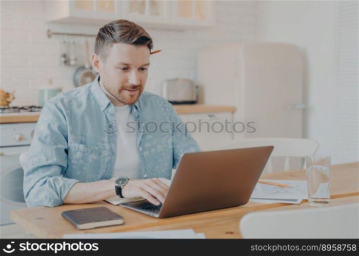 Focused young man freelancer using laptop while working from home at his workplace, millennial guy typing on pc notebook and surfing in internet, looking at screen while sitting at kitchen table. Focused young man freelancer using laptop while sitting at kitchen table