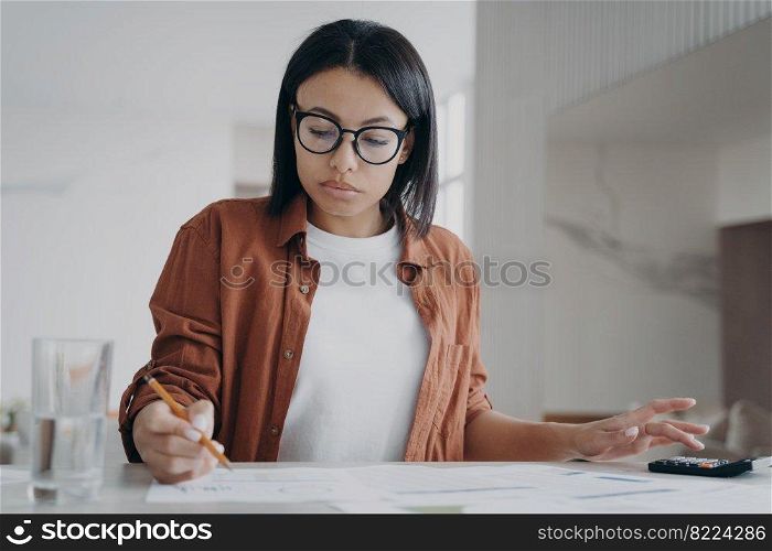 Focused young woman wearing glasses counting expenses, taxes on calculator manages family budget at home. Serious female calculates utility bills, sitting at desk. Financial management.. Woman in glasses counting expenses on calculator manages family budget at home. Financial management
