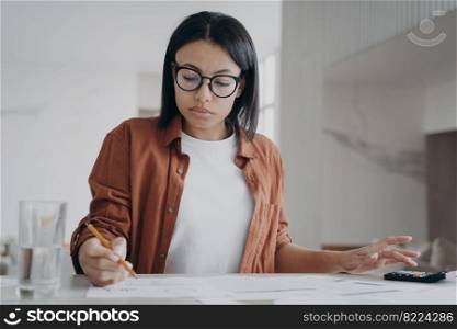 Focused young woman wearing glasses counting expenses, taxes on calculator manages family budget at home. Serious female calculates utility bills, sitting at desk. Financial management.. Woman in glasses counting expenses on calculator manages family budget at home. Financial management