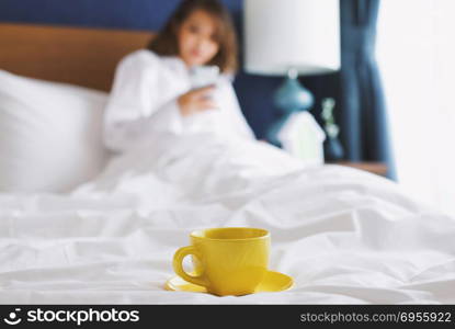 Focusing on yellow cup of coffee or tea with woman using smartphone on bed, in the morning