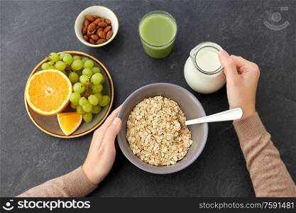 food and breakfast concept - hands of woman eating oatmeal cereals in bowl, fruits, almond nuts, glass of juice and jug with milk on slate stone table. hands with oatmeal, fruits, nuts, juice and milk