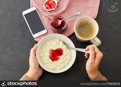 food and breakfast concept - hands of woman eating porridge in bowl, smartphone, jam in mason jar, spoon and cup of coffee on slate stone table. hands with porridge, jam, spoon, coffee and phone