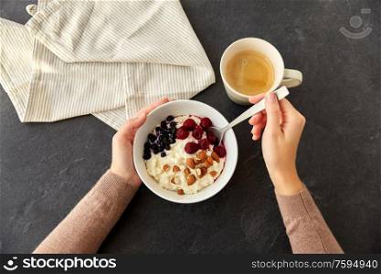 food and breakfast concept - hands of woman with spoon eating porridge in bowl with wild berries, almond nuts and cup of coffee on slate stone table. hands with porridge breakfast and cup of coffee