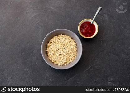 food and breakfast concept - oatmeal cereals in bowl and cup of fruit jam with spoon on slate stone table. oatmeal breakfast and cup of fruit jam with spoon