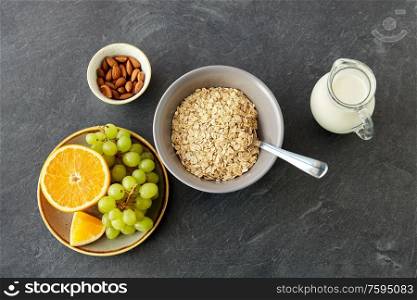 food and breakfast concept - oatmeal cereals in bowl, fruits, almond nuts and jug with milk on slate stone table. oatmeal with fruits, almond nuts and jug of milk