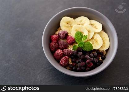 food and breakfast concept - oatmeal cereals in bowl with wild berries, banana and peppermint on slate stone table. cereal breakfast with berries, banana and mint